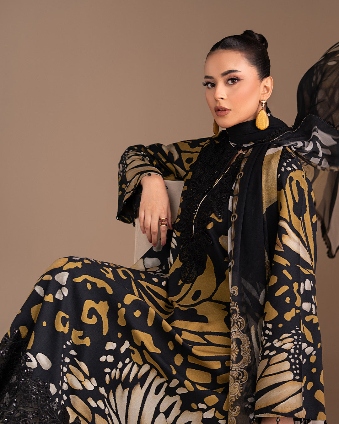 Woman wearing a black and gold patterned outfit with a scarf, sitting against a brown background.